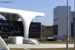 Detalhe do conjunto arquitetônico da Cidade Administrativa de Minas Gerais, em destaque estão pilares brancos, atrás deles está uma fachada toda em vidro azul escuro espelhado.