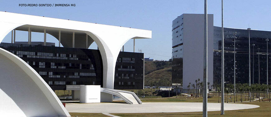 Detalhe do conjunto arquitetônico da Cidade Administrativa de Minas Gerais, em destaque estão pilares brancos, atrás deles está uma fachada toda em vidro azul escuro espelhado.