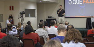 A Imagem é um registro de um auditório durante o circuito de debates sindicais. Servidoras e servidores estão sentados em fileiras, acompanhando a palestra. Ao centro, o dirigente Felipe Rodrigues fala ao microfone diante de um telão com apresentação projetada. À direita, há uma mesa de apoio com representantes sentados. O texto em destaque diz "SINJUS-MG participa de circuito de debates do Sitraemg e reforça a luta em defesa do teletrabalho". A cena registra um momento de exposição e diálogo sobre o teletrabalho no Judiciário, com identidade visual aplicada sobre a foto, destacando a participação do SINJUS-MG no evento.