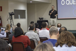 A Imagem é um registro de um auditório durante o circuito de debates sindicais. Servidoras e servidores estão sentados em fileiras, acompanhando a palestra. Ao centro, o dirigente Felipe Rodrigues fala ao microfone diante de um telão com apresentação projetada. À direita, há uma mesa de apoio com representantes sentados. O texto em destaque diz "SINJUS-MG participa de circuito de debates do Sitraemg e reforça a luta em defesa do teletrabalho". A cena registra um momento de exposição e diálogo sobre o teletrabalho no Judiciário, com identidade visual aplicada sobre a foto, destacando a participação do SINJUS-MG no evento.