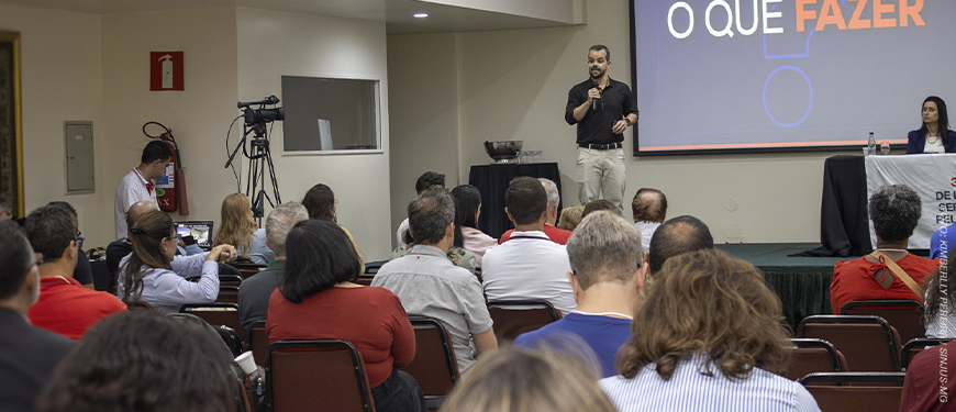 A Imagem é um registro de um auditório durante o circuito de debates sindicais. Servidoras e servidores estão sentados em fileiras, acompanhando a palestra. Ao centro, o dirigente Felipe Rodrigues fala ao microfone diante de um telão com apresentação projetada. À direita, há uma mesa de apoio com representantes sentados. O texto em destaque diz "SINJUS-MG participa de circuito de debates do Sitraemg e reforça a luta em defesa do teletrabalho". A cena registra um momento de exposição e diálogo sobre o teletrabalho no Judiciário, com identidade visual aplicada sobre a foto, destacando a participação do SINJUS-MG no evento.