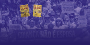 Imagem com fundo em tom roxo, mostrando uma multidão em manifestação de rua. As pessoas estão agrupadas, muitas usando roupas claras e algumas com chapéus e óculos de sol, sugerindo um ato ao ar livre durante o dia. No centro da imagem, duas mulheres em primeiro plano seguram cartazes amarelos com a frase: “Nos querem caladas, mas seguimos em luta”. Ao redor, outras pessoas também participam do protesto, algumas com expressões sérias e engajadas.