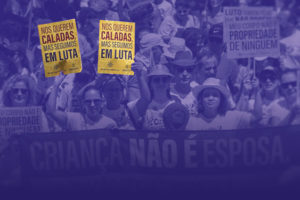 Imagem com fundo em tom roxo, mostrando uma multidão em manifestação de rua. As pessoas estão agrupadas, muitas usando roupas claras e algumas com chapéus e óculos de sol, sugerindo um ato ao ar livre durante o dia. No centro da imagem, duas mulheres em primeiro plano seguram cartazes amarelos com a frase: “Nos querem caladas, mas seguimos em luta”. Ao redor, outras pessoas também participam do protesto, algumas com expressões sérias e engajadas.