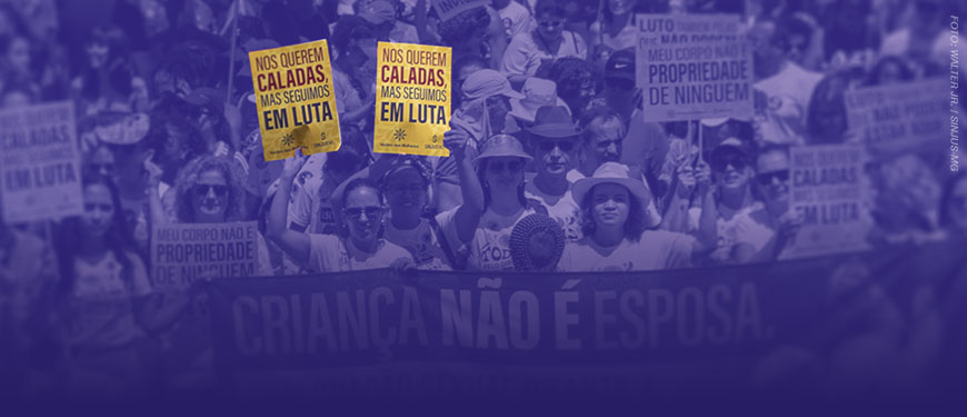 Imagem com fundo em tom roxo, mostrando uma multidão em manifestação de rua. As pessoas estão agrupadas, muitas usando roupas claras e algumas com chapéus e óculos de sol, sugerindo um ato ao ar livre durante o dia. No centro da imagem, duas mulheres em primeiro plano seguram cartazes amarelos com a frase: “Nos querem caladas, mas seguimos em luta”. Ao redor, outras pessoas também participam do protesto, algumas com expressões sérias e engajadas.