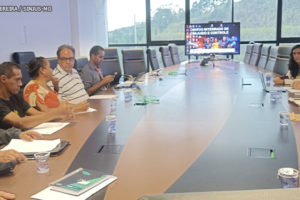 Registro de uma reunião do Conselho de Beneficiários do IPSEMG com a diretoria do hospital, na foto há pessoas diversas sentadas em uma grande mesa de reuniões, elas se dividem em dois grupos que ocupam posições opostas na mesa. Representando o SINJUS-MG está o diretor de Assuntos sociais, Culturais e Saúde, Jonas Araújo, um homem negro, sem barba, com cabelos pretos e curtos, ele veste uma camisa preta.