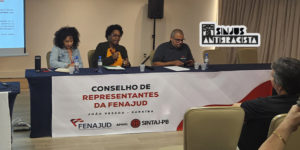 A imagem mostra duas mulheres negras sentadas à mesa durante o Conselho de Representantes da Fenajud. Cleonice Amorim, do SINJUS Antirracista, fala ao microfone enquanto a outra participante ouve atentamente. Ambas participam de um painel que debate o racismo institucional no Judiciário, reforçando o compromisso com a equidade racial nas estruturas de poder. Ao fundo, vê-se o telão com o tema do evento e a identidade visual da Fenajud.