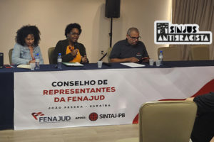 A imagem mostra duas mulheres negras sentadas à mesa durante o Conselho de Representantes da Fenajud. Cleonice Amorim, do SINJUS Antirracista, fala ao microfone enquanto a outra participante ouve atentamente. Ambas participam de um painel que debate o racismo institucional no Judiciário, reforçando o compromisso com a equidade racial nas estruturas de poder. Ao fundo, vê-se o telão com o tema do evento e a identidade visual da Fenajud.