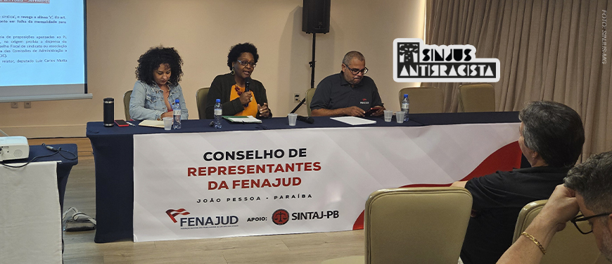 A imagem mostra duas mulheres negras sentadas à mesa durante o Conselho de Representantes da Fenajud. Cleonice Amorim, do SINJUS Antirracista, fala ao microfone enquanto a outra participante ouve atentamente. Ambas participam de um painel que debate o racismo institucional no Judiciário, reforçando o compromisso com a equidade racial nas estruturas de poder. Ao fundo, vê-se o telão com o tema do evento e a identidade visual da Fenajud.