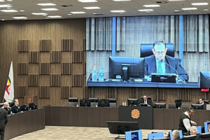 A imagem mostra uma sessão do Órgão Especial do TJMG, onde foi aprovada a proposta orçamentária do Tribunal para 2026. O registro destaca a formalidade da reunião, com os(as) desembargadores(as) em plenário e a presença de assessores(as) e servidores(as) de apoio, enquanto um telão exibe em destaque o presidente da sessão.