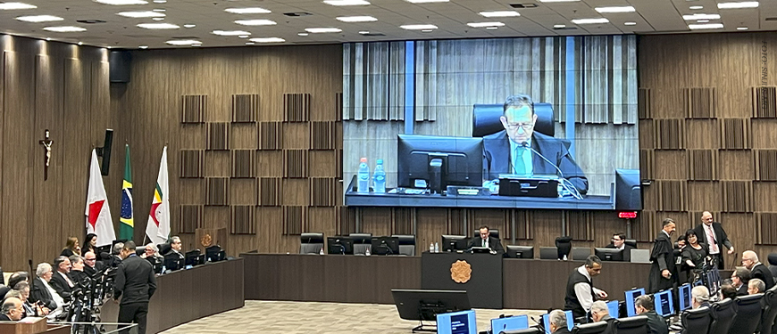 A imagem mostra uma sessão do Órgão Especial do TJMG, onde foi aprovada a proposta orçamentária do Tribunal para 2026. O registro destaca a formalidade da reunião, com os(as) desembargadores(as) em plenário e a presença de assessores(as) e servidores(as) de apoio, enquanto um telão exibe em destaque o presidente da sessão.