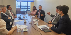 Registro de uma reunião do Grupo de Trabalho da Jornada de 8 horas, em uma sala do TJMG com mesa retangular de madeira e sete pessoas sentadas debatendo. Estão presentes representantes do TJMG e o diretor de Assuntos Jurídicos do SINJUS-MG, Felipe Rodrigues, que gesticula enquanto fala. Ao fundo, uma tela exibe um documento com anotações.