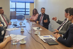 Registro de uma reunião do Grupo de Trabalho da Jornada de 8 horas, em uma sala do TJMG com mesa retangular de madeira e sete pessoas sentadas debatendo. Estão presentes representantes do TJMG e o diretor de Assuntos Jurídicos do SINJUS-MG, Felipe Rodrigues, que gesticula enquanto fala. Ao fundo, uma tela exibe um documento com anotações.