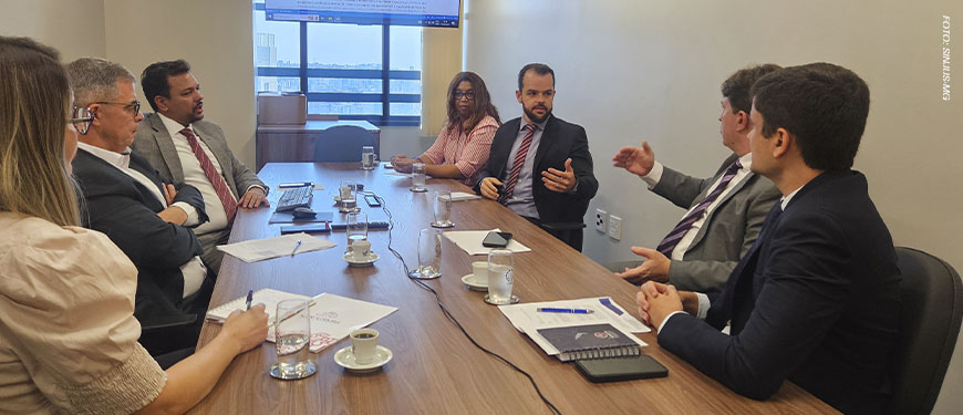 Registro de uma reunião do Grupo de Trabalho da Jornada de 8 horas, em uma sala do TJMG com mesa retangular de madeira e sete pessoas sentadas debatendo. Estão presentes representantes do TJMG e o diretor de Assuntos Jurídicos do SINJUS-MG, Felipe Rodrigues, que gesticula enquanto fala. Ao fundo, uma tela exibe um documento com anotações.
