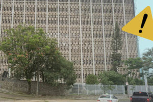 Foto diurna da fachada lateral do edifício situado à Praça Milton Campos, feita de baixo para cima. Sobre ela, é aplicada a figura de uma placa de atenção indicando alerta.