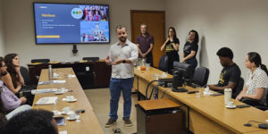 imagem mostra uma fotografia de uma reunião no Tribunal de Justiça Militar de Minas Gerais (TJMMG), realizada em uma sala ampla e iluminada. Ao centro, o diretor do SINJUS, Felipe Rodrigues, está de pé, falando aos novos servidores sentados em formato de “U”. No telão ao fundo, aparece um slide com o título “Núcleos”.