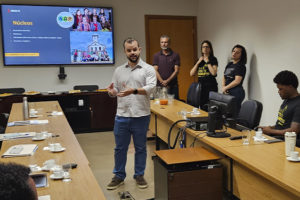 imagem mostra uma fotografia de uma reunião no Tribunal de Justiça Militar de Minas Gerais (TJMMG), realizada em uma sala ampla e iluminada. Ao centro, o diretor do SINJUS, Felipe Rodrigues, está de pé, falando aos novos servidores sentados em formato de “U”. No telão ao fundo, aparece um slide com o título “Núcleos”.