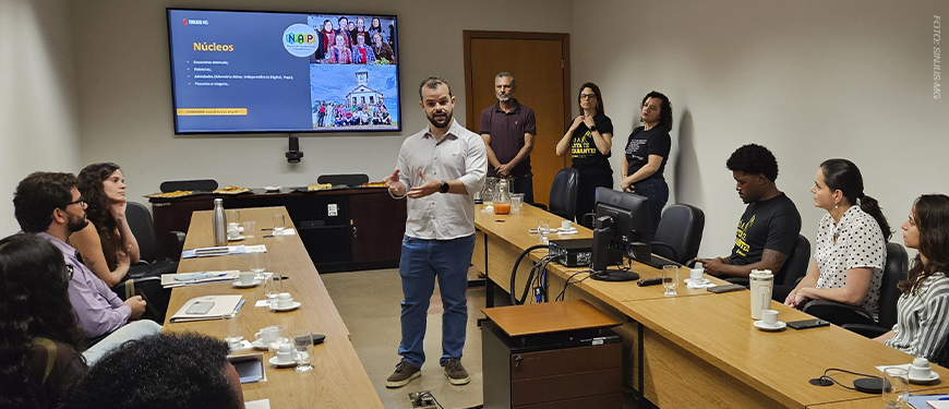 imagem mostra uma fotografia de uma reunião no Tribunal de Justiça Militar de Minas Gerais (TJMMG), realizada em uma sala ampla e iluminada. Ao centro, o diretor do SINJUS, Felipe Rodrigues, está de pé, falando aos novos servidores sentados em formato de “U”. No telão ao fundo, aparece um slide com o título “Núcleos”.
