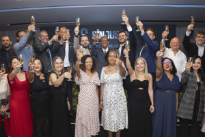 A imagem mostra os integrantes da nova diretoria do SINJUS-MG reunida em um evento de posse, todos vestidos formalmente erguendo taças em um brinde coletivo. No topo lê-se “Unidade”. O texto principal informa: “SINJUS-MG inicia novo ciclo de lutas com posse da gestão 2026-2029”. A cena representa a cerimônia de posse da Diretoria Colegiada e do Conselho Fiscal do sindicato, marcada pela celebração, renovação do compromisso com a categoria e defesa dos direitos das servidoras e dos servidores do Judiciário mineiro.