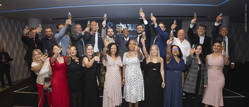 A imagem mostra os integrantes da nova diretoria do SINJUS-MG reunida em um evento de posse, todos vestidos formalmente erguendo taças em um brinde coletivo. No topo lê-se “Unidade”. O texto principal informa: “SINJUS-MG inicia novo ciclo de lutas com posse da gestão 2026-2029”. A cena representa a cerimônia de posse da Diretoria Colegiada e do Conselho Fiscal do sindicato, marcada pela celebração, renovação do compromisso com a categoria e defesa dos direitos das servidoras e dos servidores do Judiciário mineiro.
