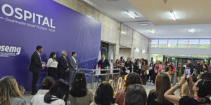 A imagem mostra ao fundo uma foto de solenidade de reinauguração da ala do Hospital Governador Israel Pinheiro, do Ipsemg. No palco, autoridades aparecem diante de um grande painel roxo com a identificação do hospital, enquanto o público acompanha o evento.