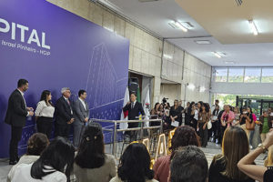 A imagem mostra ao fundo uma foto de solenidade de reinauguração da ala do Hospital Governador Israel Pinheiro, do Ipsemg. No palco, autoridades aparecem diante de um grande painel roxo com a identificação do hospital, enquanto o público acompanha o evento.