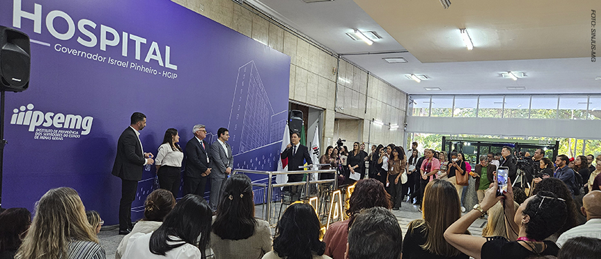 A imagem mostra ao fundo uma foto de solenidade de reinauguração da ala do Hospital Governador Israel Pinheiro, do Ipsemg. No palco, autoridades aparecem diante de um grande painel roxo com a identificação do hospital, enquanto o público acompanha o evento.