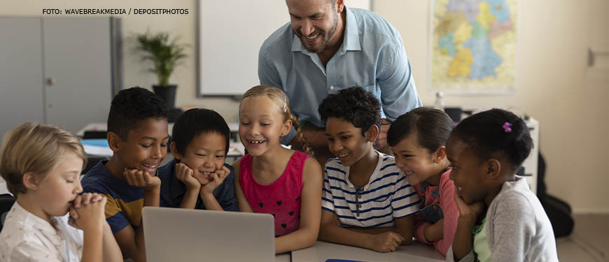 Imagem Acessível: Em um ambiente escolar com um mapa ao fundo, há um homem branco adulto está com um grupo de crianças de diversas etnias, há brancos, negros e asiáticos. O homem sorri enquanto apresenta um conteúdo em computador para as crianças.