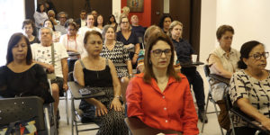 Imagem Acessível: A imagem mostra uma sala iluminada, com diversos integrantes do NAP sentadas em cadeiras organizadas em fileiras. A maioria mantém os olhos fechados e postura relaxada, participando de uma prática de respiração consciente. No topo da imagem, aparece o título: “NAP promove encontro sobre respiração consciente e qualidade de vida”.