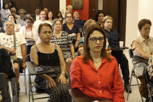 Imagem Acessível: A imagem mostra uma sala iluminada, com diversos integrantes do NAP sentadas em cadeiras organizadas em fileiras. A maioria mantém os olhos fechados e postura relaxada, participando de uma prática de respiração consciente. No topo da imagem, aparece o título: “NAP promove encontro sobre respiração consciente e qualidade de vida”.