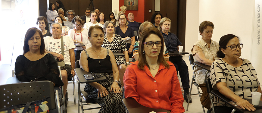 Imagem Acessível: A imagem mostra uma sala iluminada, com diversos integrantes do NAP sentadas em cadeiras organizadas em fileiras. A maioria mantém os olhos fechados e postura relaxada, participando de uma prática de respiração consciente. No topo da imagem, aparece o título: “NAP promove encontro sobre respiração consciente e qualidade de vida”.