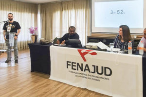 Foto do I Seminário Internacional da FENAJUD onde o diretor de Finanças do SINJUS-MG, Felipe Rodrigues, se encontra à esquerda da foto, de pé em frente a um microfone ao seu lado direito uma mesa composta pela presidente da Internacional de Serviços Públicos (ISP), Denise Motta Dau; o presidente da Confederação Latino-americana dos Trabalhadores do Poder Judiciário (CLTPJ), Ariel Pringles e o membro do Conselho Executivo da Confederação Sindical das Américas (CSA), o advogado Marcelo Di Stefano. Sobre a mesa uma bandeira branca com a logo da FENAJUD.