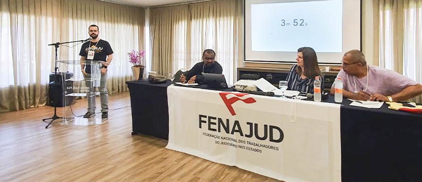 Foto do I Seminário Internacional da FENAJUD onde o diretor de Finanças do SINJUS-MG, Felipe Rodrigues, se encontra à esquerda da foto, de pé em frente a um microfone ao seu lado direito uma mesa composta pela presidente da Internacional de Serviços Públicos (ISP), Denise Motta Dau; o presidente da Confederação Latino-americana dos Trabalhadores do Poder Judiciário (CLTPJ), Ariel Pringles e o membro do Conselho Executivo da Confederação Sindical das Américas (CSA), o advogado Marcelo Di Stefano. Sobre a mesa uma bandeira branca com a logo da FENAJUD.