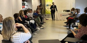 Imagem Acessível: Encontro promovido pelo NAP em uma sala de reuniões, com participantes sentados em semicírculo enquanto o palestrante em pé apresenta orientações. Ao fundo, há um banner do NAP e uma mesa com a marca SINJUS-MG.Conteúdo textual: Prevenção - NAP promove encontro e reforça cuidados para evitar golpes digitais