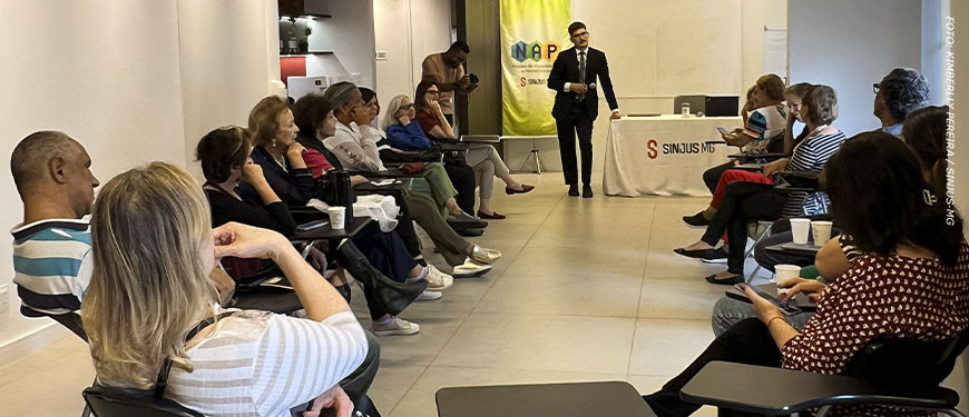 Imagem Acessível: Encontro promovido pelo NAP em uma sala de reuniões, com participantes sentados em semicírculo enquanto o palestrante em pé apresenta orientações. Ao fundo, há um banner do NAP e uma mesa com a marca SINJUS-MG.Conteúdo textual: Prevenção - NAP promove encontro e reforça cuidados para evitar golpes digitais