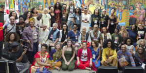 Foto de um grande grupo de participantes do Seminário Internacional sobre reparações aos povos negros, reunidos em um auditório diante de um mural colorido com cenas culturais. As pessoas estão sorrindo e posando para a foto, usando roupas diversas e coloridas. Entre os participantes estão as representantes do SINJUS-MG: Cleo Amorim, do coletivo SINJUS Antirracista e as dirigentes do Sindicato Adriana Teodoro e Patrícia Oliveira.