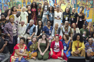Foto de um grande grupo de participantes do Seminário Internacional sobre reparações aos povos negros, reunidos em um auditório diante de um mural colorido com cenas culturais. As pessoas estão sorrindo e posando para a foto, usando roupas diversas e coloridas. Entre os participantes estão as representantes do SINJUS-MG: Cleo Amorim, do coletivo SINJUS Antirracista e as dirigentes do Sindicato Adriana Teodoro e Patrícia Oliveira.