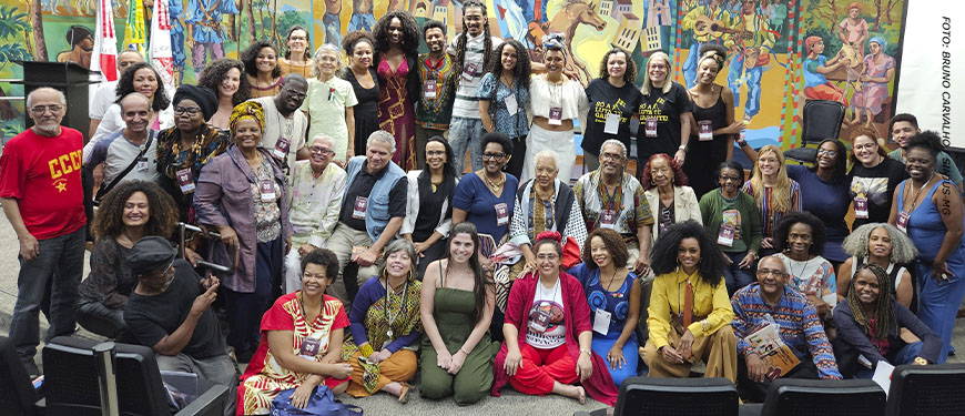 Foto de um grande grupo de participantes do Seminário Internacional sobre reparações aos povos negros, reunidos em um auditório diante de um mural colorido com cenas culturais. As pessoas estão sorrindo e posando para a foto, usando roupas diversas e coloridas. Entre os participantes estão as representantes do SINJUS-MG: Cleo Amorim, do coletivo SINJUS Antirracista e as dirigentes do Sindicato Adriana Teodoro e Patrícia Oliveira.