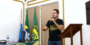 A imagem mostra o diretor de Comunicação e Imprensa do SINJUS-MG, Alexandre Gomes, falando ao microfone durante a Assembleia Geral Ordinária do Conselho de Representantes da Confederação dos Servidores Públicos do Brasil (CSPB). Ao fundo, há bandeiras do Brasil, da CSPB e de entidades sindicais. O ambiente possui paredes claras com detalhes dourados e iluminação suave.