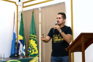 A imagem mostra o diretor de Comunicação e Imprensa do SINJUS-MG, Alexandre Gomes, falando ao microfone durante a Assembleia Geral Ordinária do Conselho de Representantes da Confederação dos Servidores Públicos do Brasil (CSPB). Ao fundo, há bandeiras do Brasil, da CSPB e de entidades sindicais. O ambiente possui paredes claras com detalhes dourados e iluminação suave.