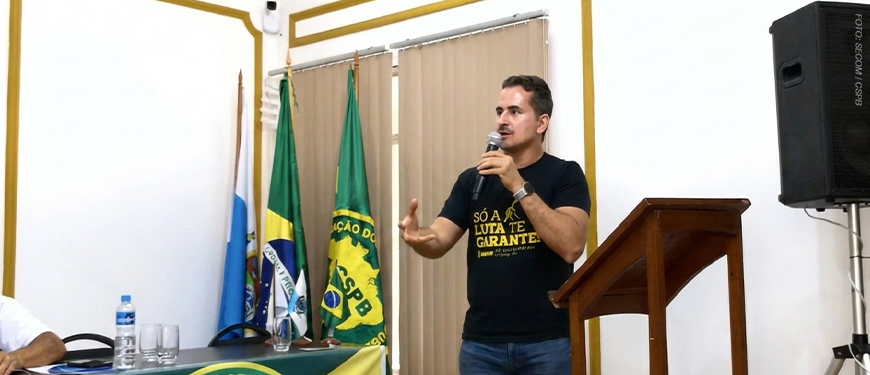 A imagem mostra o diretor de Comunicação e Imprensa do SINJUS-MG, Alexandre Gomes, falando ao microfone durante a Assembleia Geral Ordinária do Conselho de Representantes da Confederação dos Servidores Públicos do Brasil (CSPB). Ao fundo, há bandeiras do Brasil, da CSPB e de entidades sindicais. O ambiente possui paredes claras com detalhes dourados e iluminação suave.