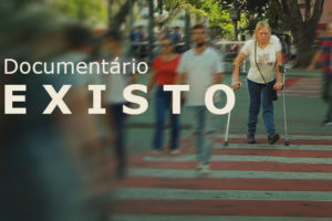 A imagem divulga o documentário “Existo”. Ao fundo, uma cena urbana com faixa de pedestres movimentada; várias pessoas aparecem desfocadas em movimento, enquanto duas mulheres com deficiência física, usando muletas, caminham com nitidez pelo centro da faixa. Sobre a imagem, os textos em destaque informam: “Documentário Existo”, data “24/4 (sexta-feira)”, horário “20h” e local “Una Cine Belas Artes”. À direita, destaca-se a mensagem “Inscrições abertas – vagas limitadas”. Na parte inferior, aparece o logotipo do SINJUS MG.