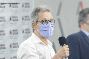 #ImagemAcessível: Foto do governador de Minas, Romeu Zema segurando um microfone (homem de pele clara, cabelos grisalhos, usando camisa social branca, óculos de grau com a armação preta e máscara na cor azul).