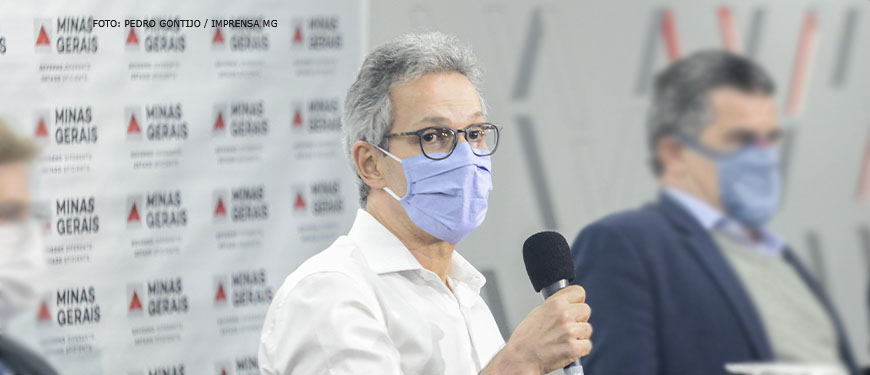 #ImagemAcessível: Foto do governador de Minas, Romeu Zema segurando um microfone (homem de pele clara, cabelos grisalhos, usando camisa social branca, óculos de grau com a armação preta e máscara na cor azul).