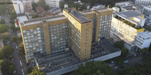 Fotografia diurna, panorâmica vista de cima da unidade IPSEMG, um prédio na cor marrom claro, com janelas, em uma rua arborizada e movimentada por automóveis, em torno da unidade se vê prédios e casas.