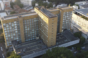Fotografia diurna, panorâmica vista de cima da unidade IPSEMG, um prédio na cor marrom claro, com janelas, em uma rua arborizada e movimentada por automóveis, em torno da unidade se vê prédios e casas.