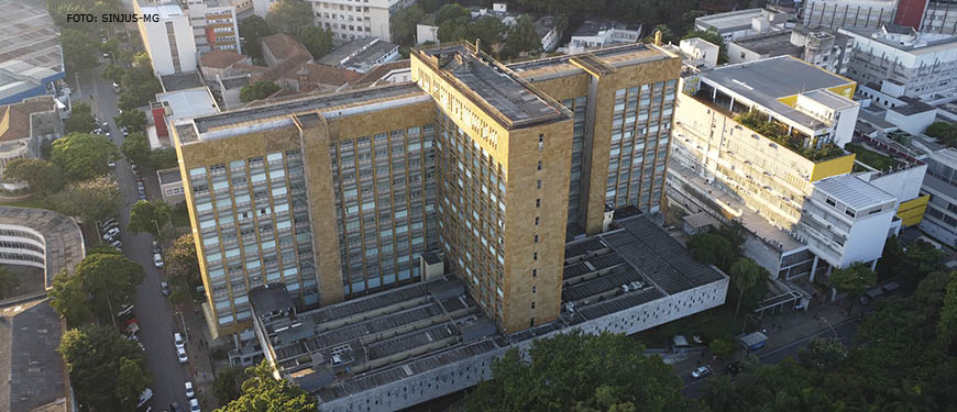 Fotografia diurna, panorâmica vista de cima da unidade IPSEMG, um prédio na cor marrom claro, com janelas, em uma rua arborizada e movimentada por automóveis, em torno da unidade se vê prédios e casas.