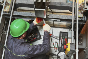 A imagem mostra um trabalhador dentro do poço de um elevador. Ele está usando um capacete verde e um uniforme de trabalho cinza. Ele está agachado sobre a estrutura do elevador, realizando manutenção. À sua volta, há várias ferramentas espalhadas, incluindo alicates, chaves de fenda e uma caixa de ferramentas. Cabos de aço grossos estão suspensos no centro da imagem, e as paredes do poço do elevador são de concreto. A iluminação é limitada, sugerindo que o local está em construção ou em manutenção.