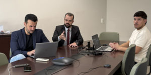 Na foto, estão Fellipe Tomanini, diretor de Assuntos Jurídicos, com cabelos curtos e escuros, barba aparada, blazer azul-marinho e camisa preta; Felipe Rodrigues, coordenador-geral do SINJUS-MG, com cabelos curtos e escuros, barba cheia, terno preto, camisa branca e gravata vinho; e Augusto Alves, advogado do Sindicato, com cabelos curtos e escuros, bigode e barba rala, vestindo camiseta branca. Os três estão sentados ao redor de uma mesa, com notebooks e equipamentos utilizados na audiência virtual.