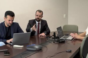 Na foto, estão Fellipe Tomanini, diretor de Assuntos Jurídicos, com cabelos curtos e escuros, barba aparada, blazer azul-marinho e camisa preta; Felipe Rodrigues, coordenador-geral do SINJUS-MG, com cabelos curtos e escuros, barba cheia, terno preto, camisa branca e gravata vinho; e Augusto Alves, advogado do Sindicato, com cabelos curtos e escuros, bigode e barba rala, vestindo camiseta branca. Os três estão sentados ao redor de uma mesa, com notebooks e equipamentos utilizados na audiência virtual.