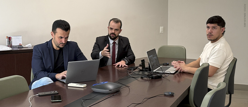 Na foto, estão Fellipe Tomanini, diretor de Assuntos Jurídicos, com cabelos curtos e escuros, barba aparada, blazer azul-marinho e camisa preta; Felipe Rodrigues, coordenador-geral do SINJUS-MG, com cabelos curtos e escuros, barba cheia, terno preto, camisa branca e gravata vinho; e Augusto Alves, advogado do Sindicato, com cabelos curtos e escuros, bigode e barba rala, vestindo camiseta branca. Os três estão sentados ao redor de uma mesa, com notebooks e equipamentos utilizados na audiência virtual.